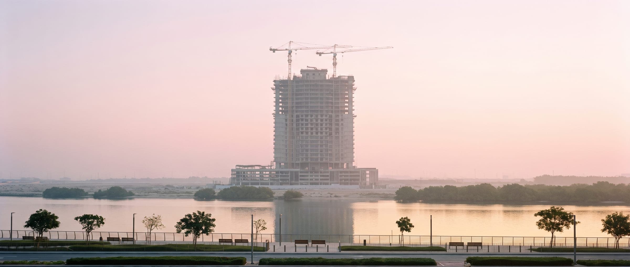 Residential tower under construction at dawn beside the creek, cranes softened by haze