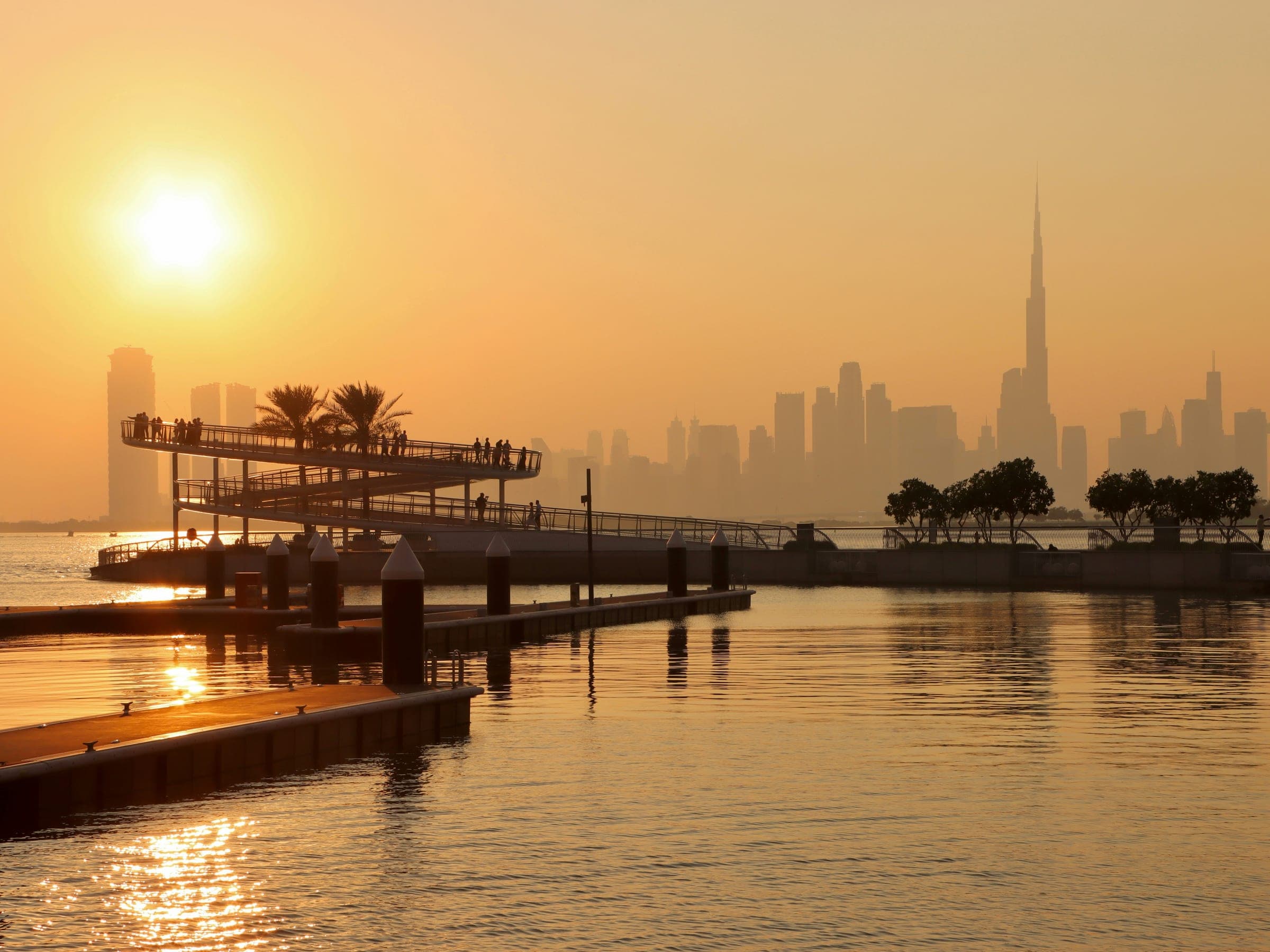 Photograph of the spiral viewing platform and marina pontoons at Dubai Creek Harbour at sunset, with the Downtown Dubai skyline in haze across the water