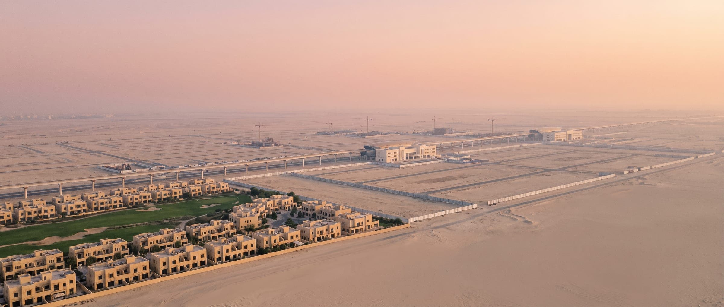 Aerial view of the Dubai South master plan at dawn: a small cluster of finished townhouses and a golf course, an elevated metro line, and graded empty plots to the horizon