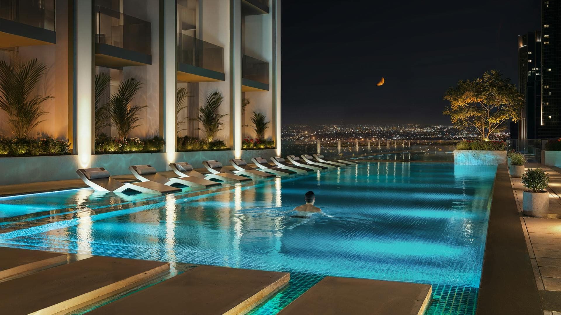 Render of the pool at Binghatti Etherea at night, a row of loungers along the lit water beside planted terraces with city lights beyond